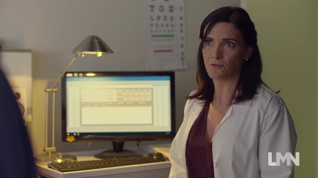 A woman in a white coat stands in a doctor's office, speaking to someone out of frame. The computer screen behind her displays a spreadsheet, and a Lifetime Movies logo is visible in the corner.
