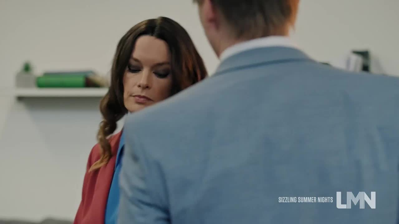 A woman in a red jacket and blue shirt looks down, her face serious. A man in a light blue suit stands in front of her, his back to the camera.
