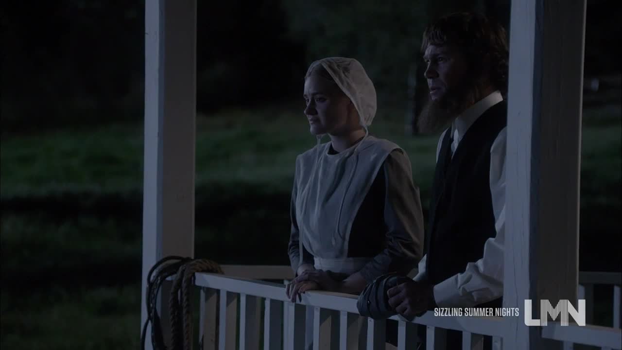 A man and woman, dressed in plain clothing, stand on a porch looking out at the dark. They lean on the white railing, their faces somber.
