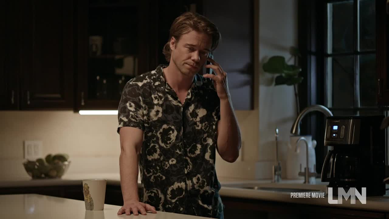 A man in a patterned shirt stands in a kitchen, holding a phone to his ear. He leans against a countertop, looking concerned as he listens to the call.
