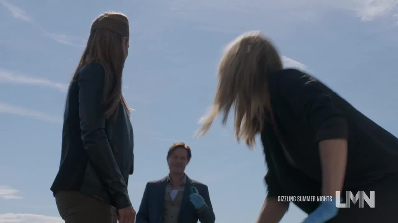 A woman with blonde hair bends over, her head angled down. A man in a suit and blue gloves stands between the two women, watching.
