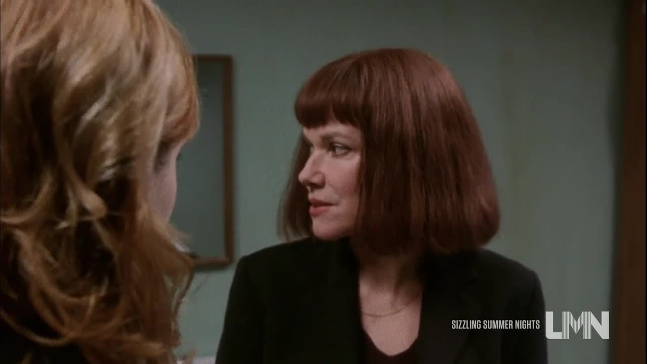 A woman with a bob haircut and a dark blazer is looking at someone off-screen. Her expression is serious, and she seems to be listening intently.
