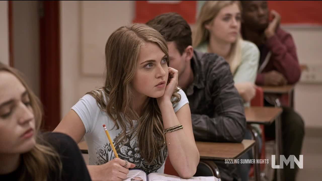 A young woman in a light blue t-shirt rests her chin on her hand, staring off to the side.  She holds a pencil over an open notebook, seemingly lost in thought in a classroom setting.
