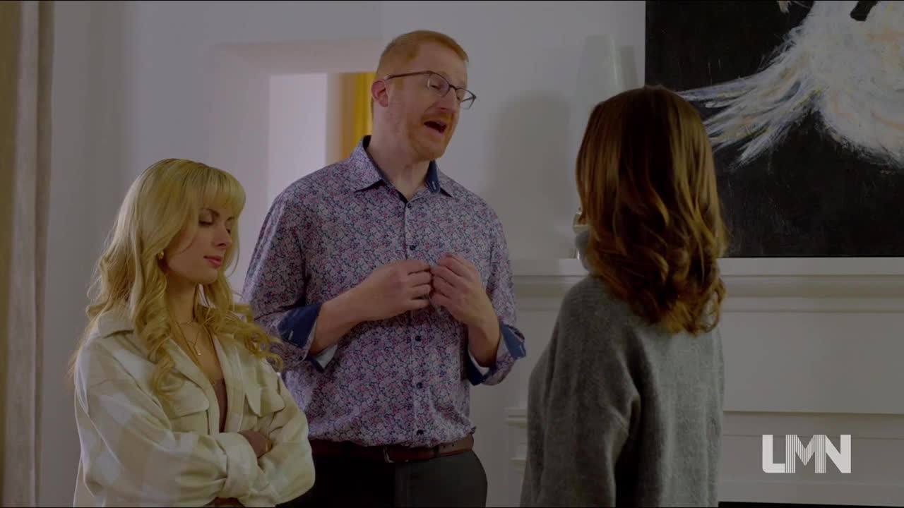 A man in a floral shirt gestures animatedly, speaking to a woman with her back turned. Another woman, blonde and in a cream-colored jacket, looks on with a guarded expression.
