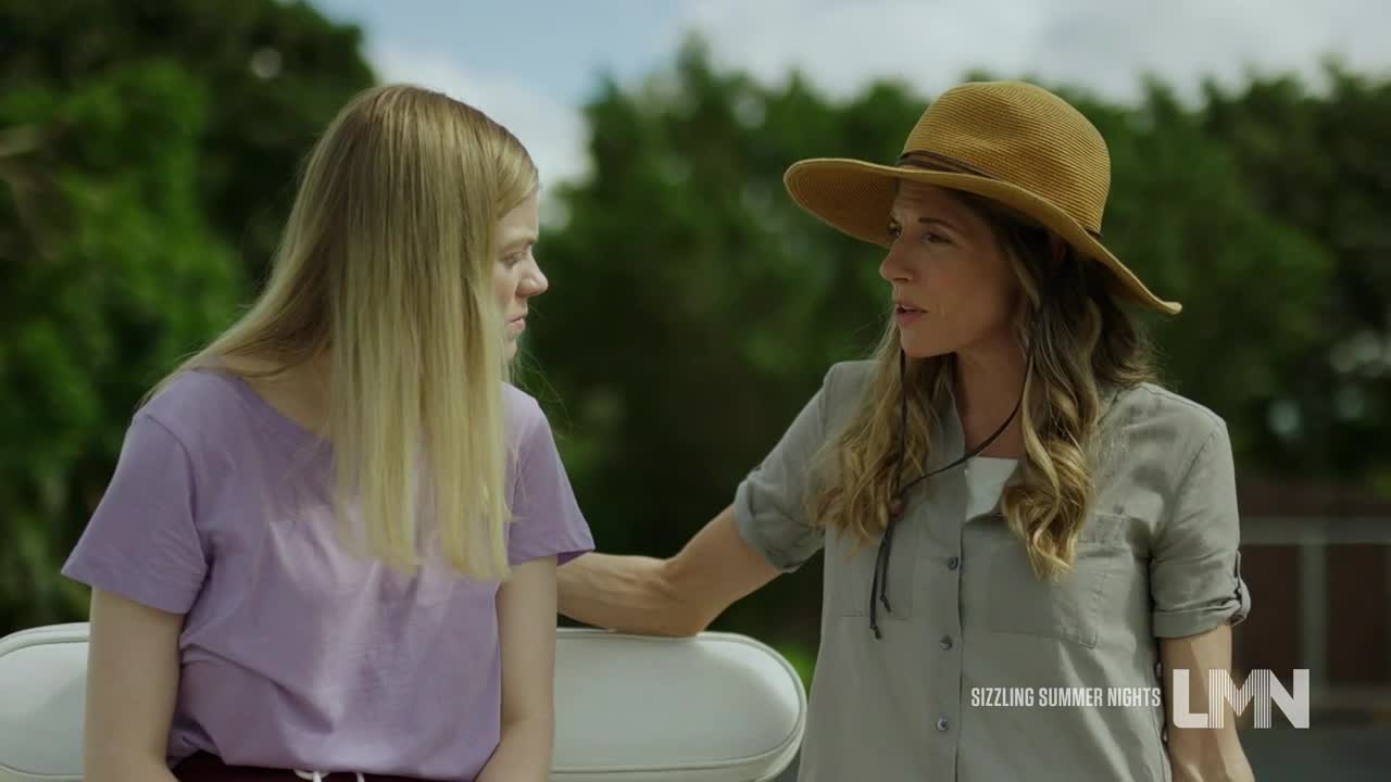 A woman in a wide-brimmed hat leans toward a younger woman, her hand resting gently on her arm. The other woman, wearing a purple shirt, looks down, listening.
