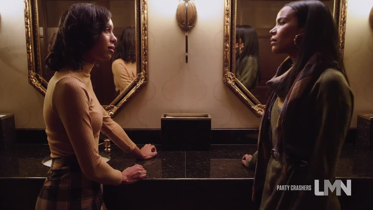 Two women stand facing each other in a bathroom, their reflections framed in ornate mirrors. One wears a tan turtleneck and plaid skirt, the other a green coat, both leaning on the countertop.
