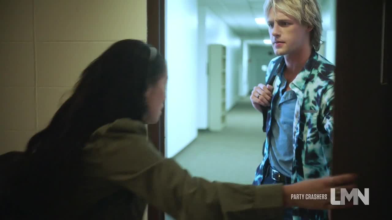 A woman, framed in a doorway, seems to be speaking to a man in a tie-dye shirt. He stands in a long, empty hallway, a backpack slung over his shoulder.
