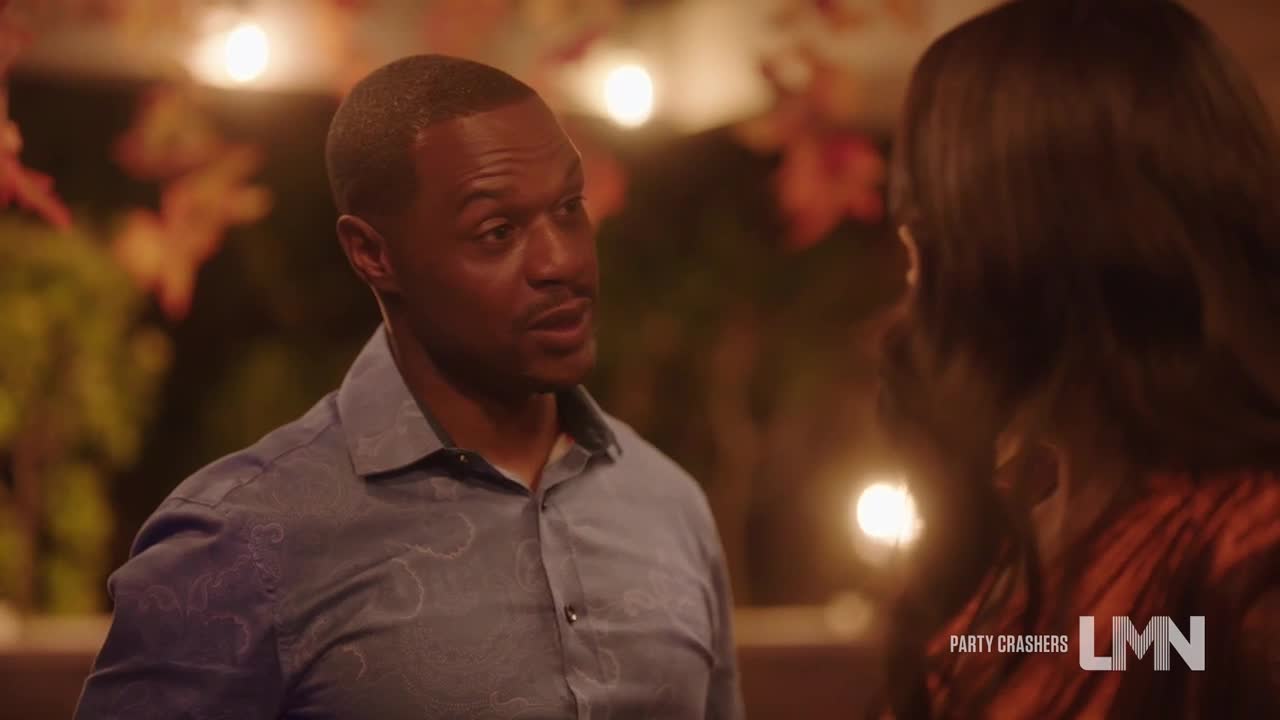 A man in a blue patterned shirt is talking, his expression earnest. The woman he's addressing is mostly out of view, wearing an orange top, and the scene is set outdoors, possibly at a party.
