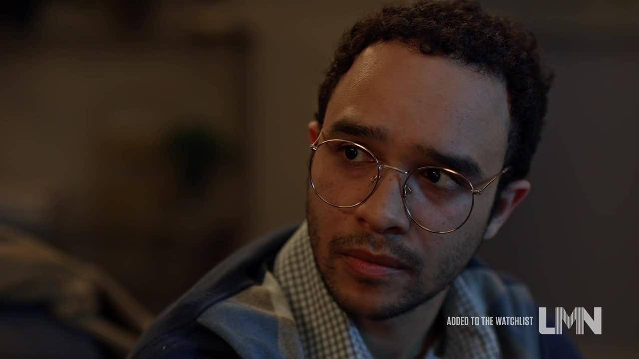 He's looking off to the side, his gaze thoughtful behind round, gold-rimmed glasses. The scene, a Lifetime movie, appears to be indoors, with a soft light illuminating his face.
