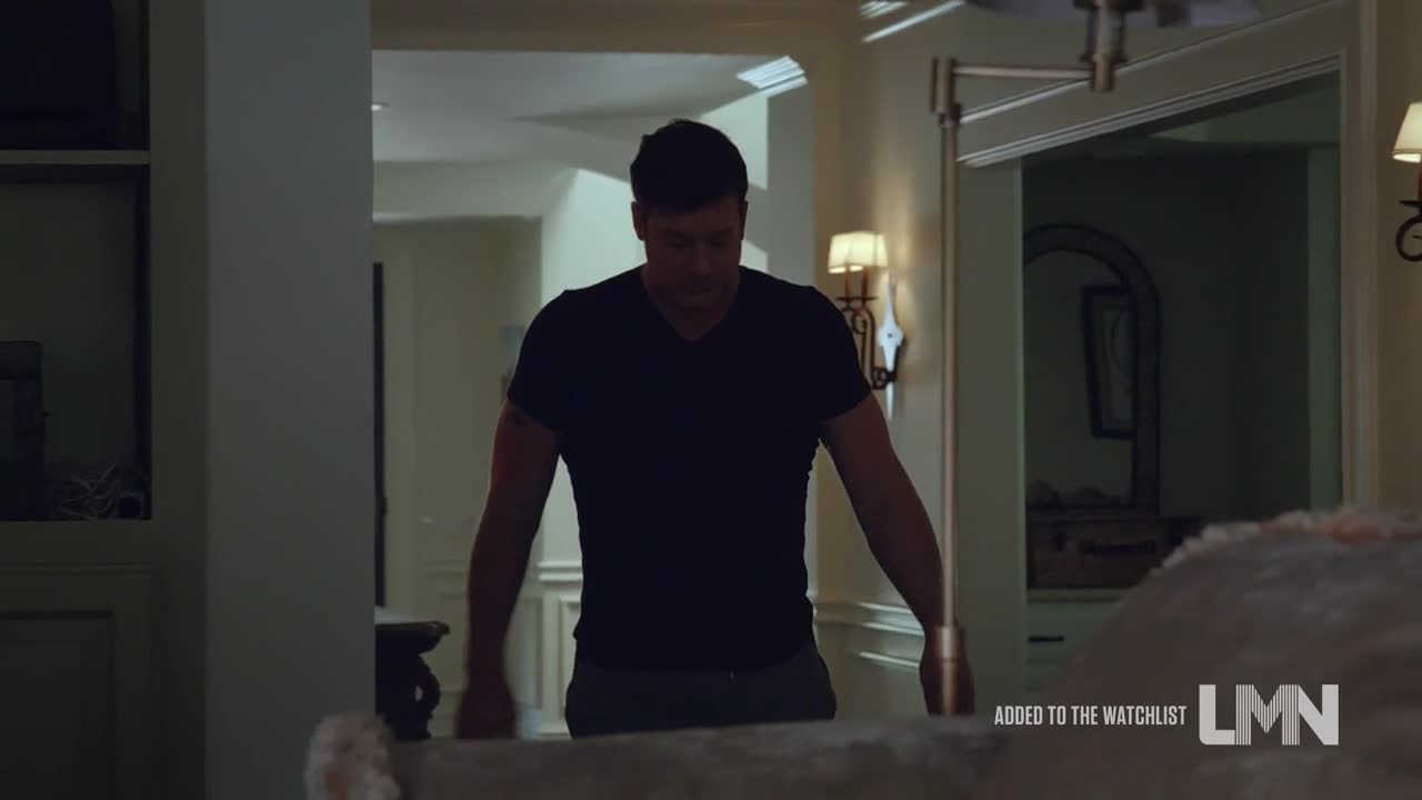 A man in a dark t-shirt walks toward the camera, his expression unreadable. He's moving through a well-lit hallway, the doorway behind him leading to another room.
