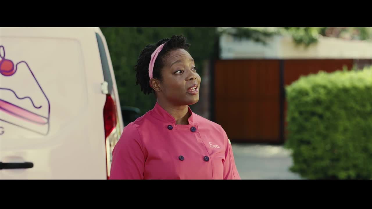 A woman in a pink chef's coat stands beside a white van, her expression animated. She's looking up and to the right, seemingly engaged in a conversation.
