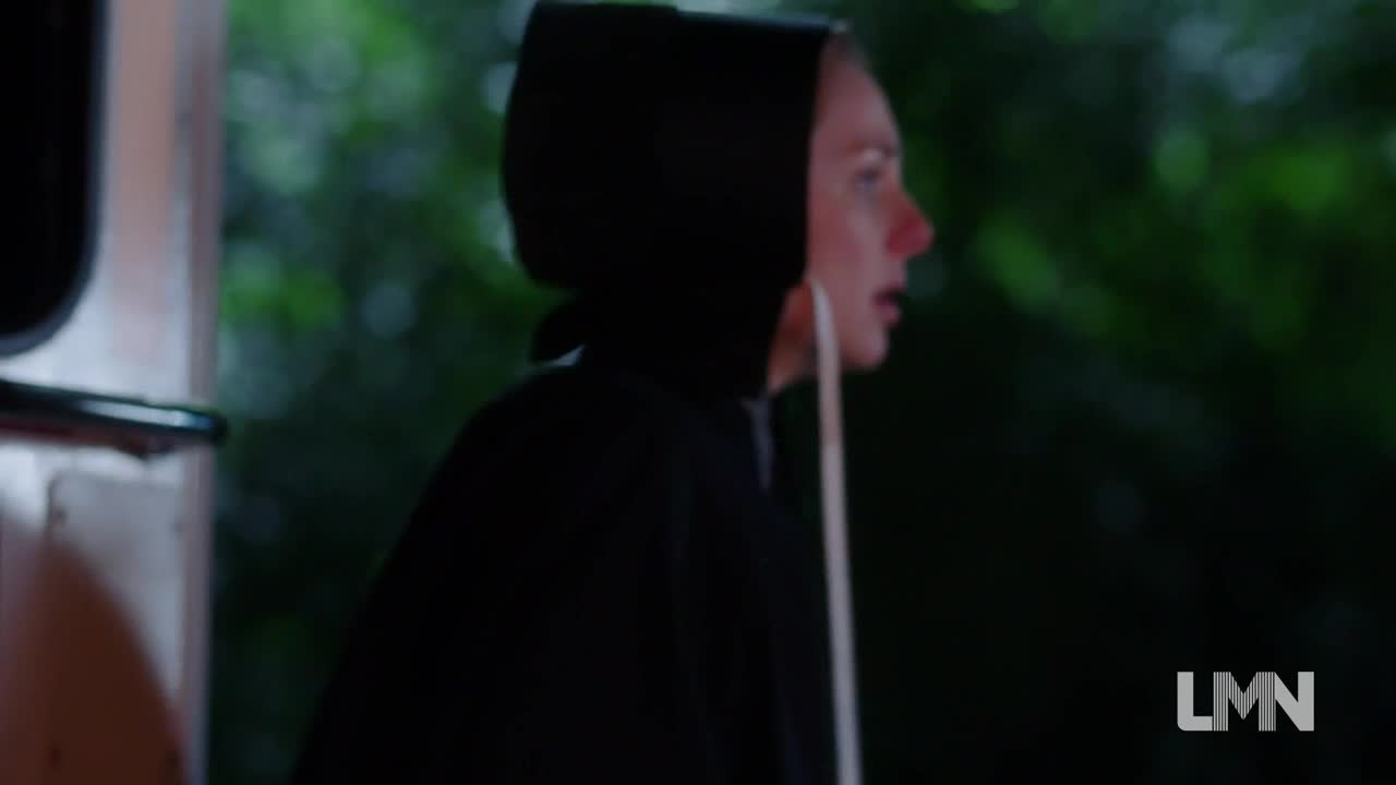 A woman in a black hood stares intently to her right, her face illuminated by an unseen light source. A white object hangs down in front of her.
