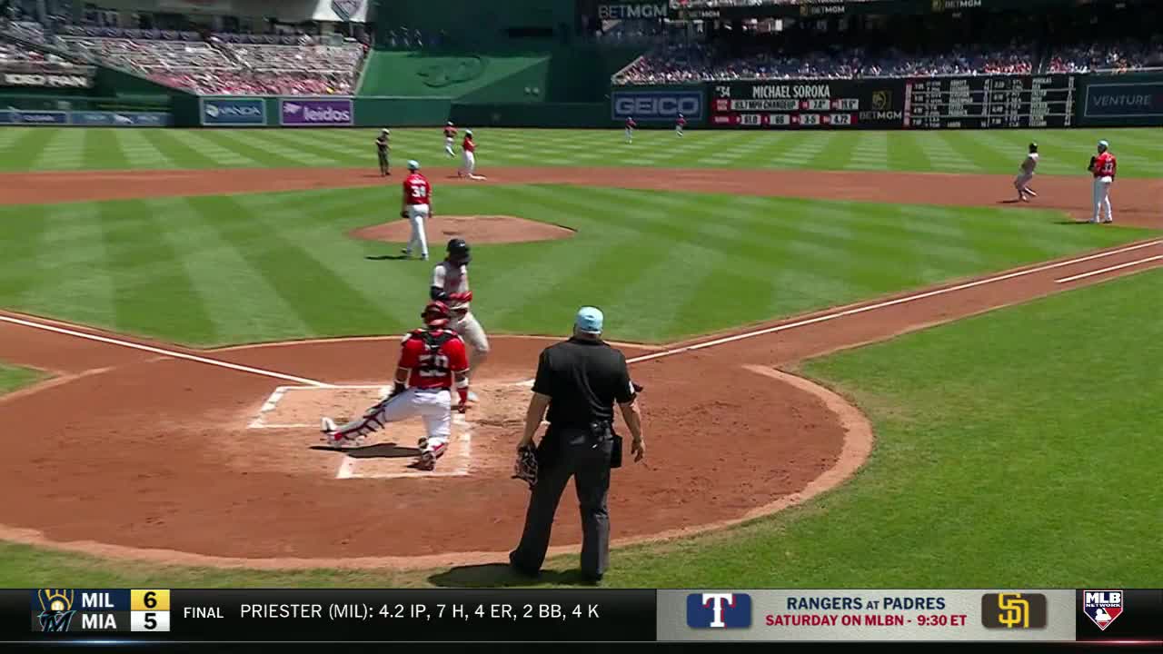 The catcher in red gear is crouched low, watching as the runner heads toward first base. The umpire stands behind the catcher, ready to make the call.
