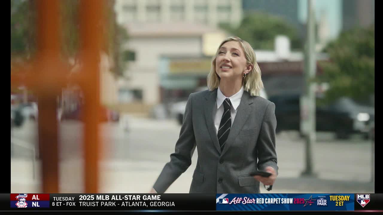 A woman in a suit, tie, and earrings looks upward with a smile, holding a phone in her hand. The MLB Network graphic at the bottom indicates a broadcast about the All-Star Game.
