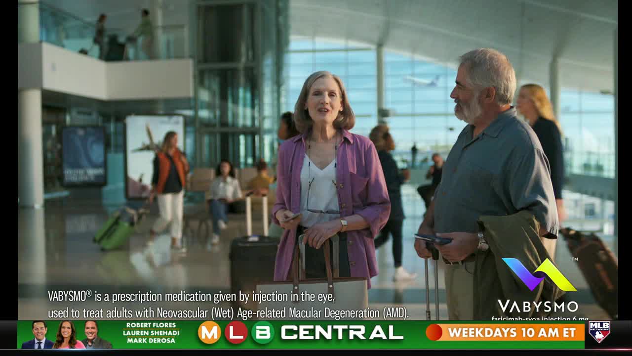 A couple stands in an airport, smiling and looking toward the camera. The MLB Network logo is visible at the bottom, and a commercial for VABYSMO is playing.
