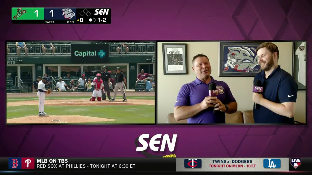 On the field, a pitcher is poised, ready to throw, while the catcher kneels behind home plate. Across the screen, two men on the MLB Network are holding microphones, smiling and talking.
