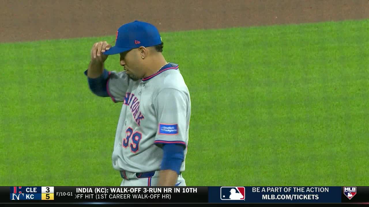 The New York Mets player, wearing number 39, is standing on the field, adjusting his cap. He looks down, as the MLB Network broadcast shows the score of the game.
