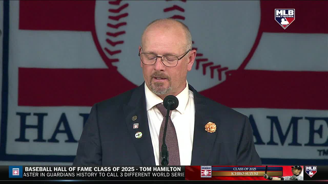 A man in a suit stands at a podium, speaking into a microphone on MLB Network. Behind him, the Baseball Hall of Fame logo is visible.
