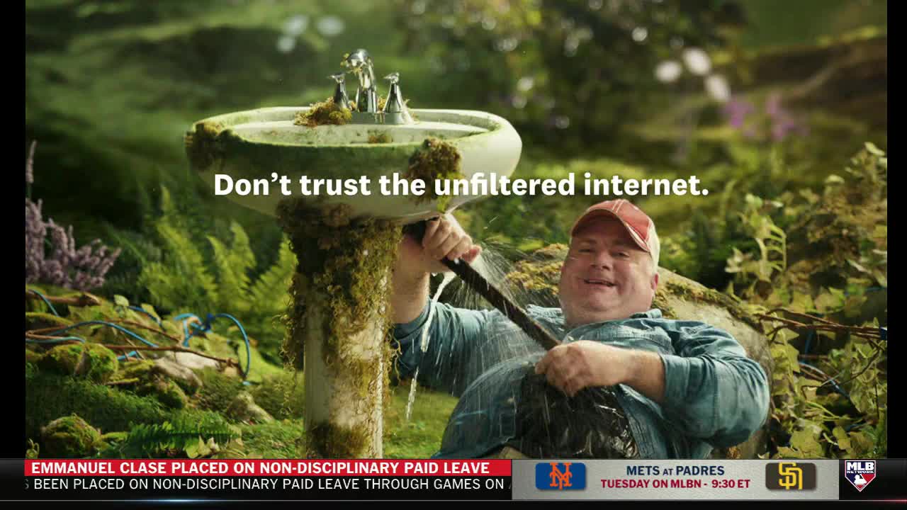 A man in a baseball cap sprays water from a hose at a moss-covered sink, set in a lush garden. The MLB Network graphic at the bottom reminds me the Mets are playing the Padres on Tuesday.
