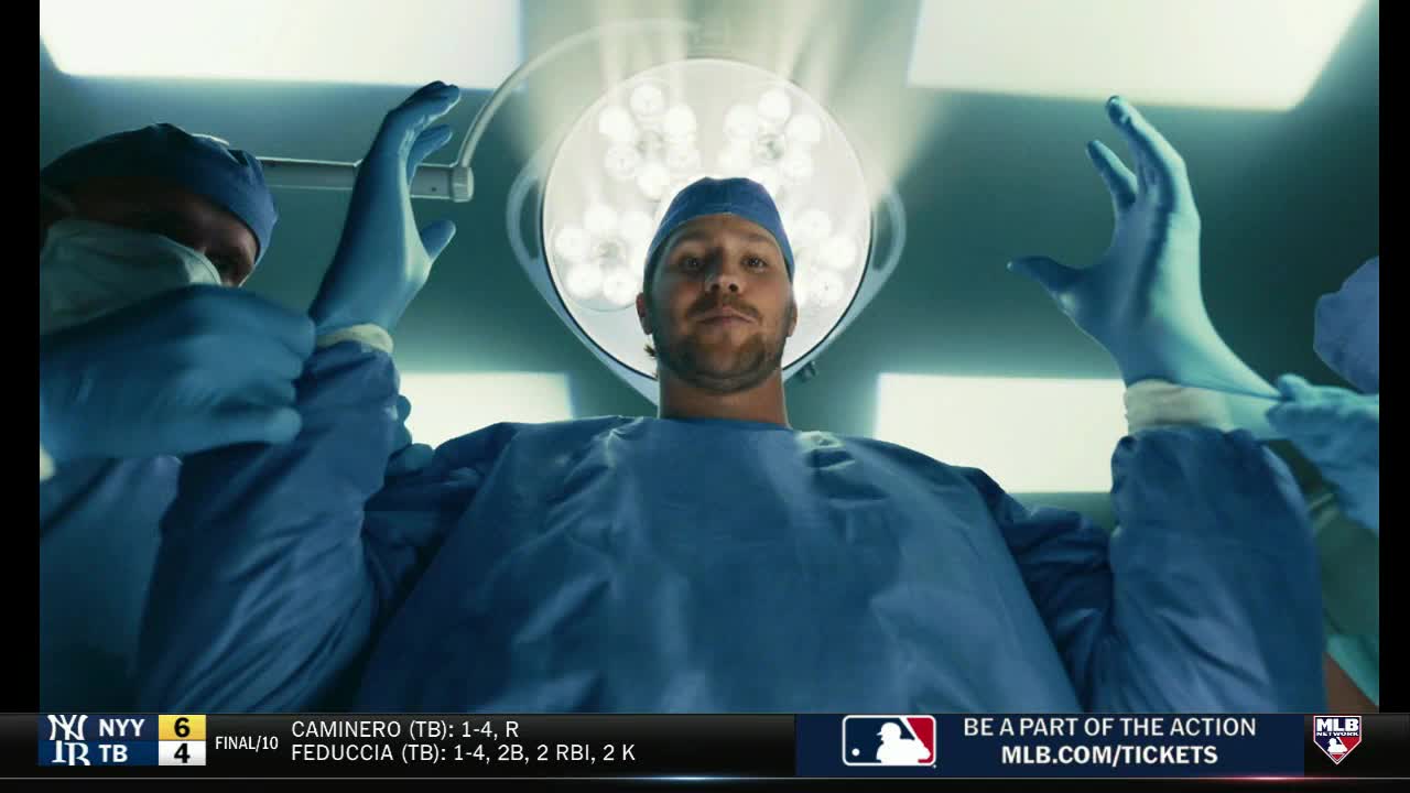 Under a bright operating room light, a baseball player in a surgical gown stares directly at the camera. Gloved hands reach in from the sides, as if preparing him for a procedure.
