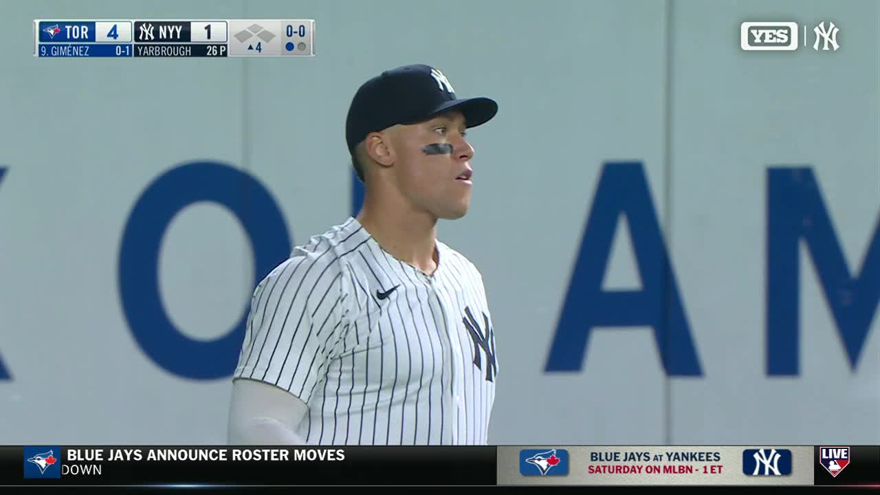 Aaron Judge, in his pinstripes, stands on the field, looking toward the action. The scoreboard shows the Yankees trailing the Blue Jays.
