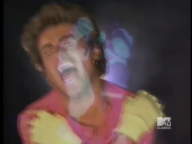 A man in a pink shirt and yellow sleeves is singing with his mouth wide open. Ghostly, multi-colored overlays ripple over him, a visual effect from a music video on MTV Classic.
