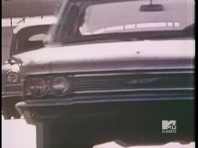 A silver car with a prominent grille is parked, its headlights reflecting the sun. The scene, reminiscent of a vintage American film, is marked by the MTV Classic logo.

