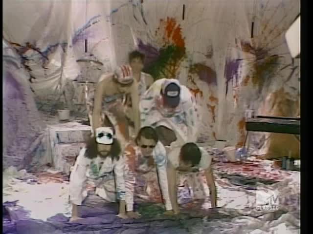 Five guys, covered in paint, are crawling around on a floor splattered with color. Behind them, a backdrop of white sheets is also covered in paint, the whole scene looking like a messy art project.
