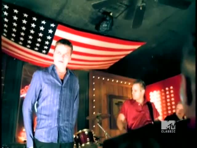 A man in a blue shirt stands center stage, while another in red plays a keyboard, both under a huge American flag. The MTV Classic logo is visible in the corner.
