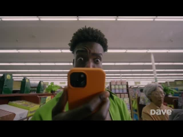 A young man, holding an orange phone, stares wide-eyed directly at the camera. He's in a brightly lit grocery store, and an older woman looks on from behind him.
