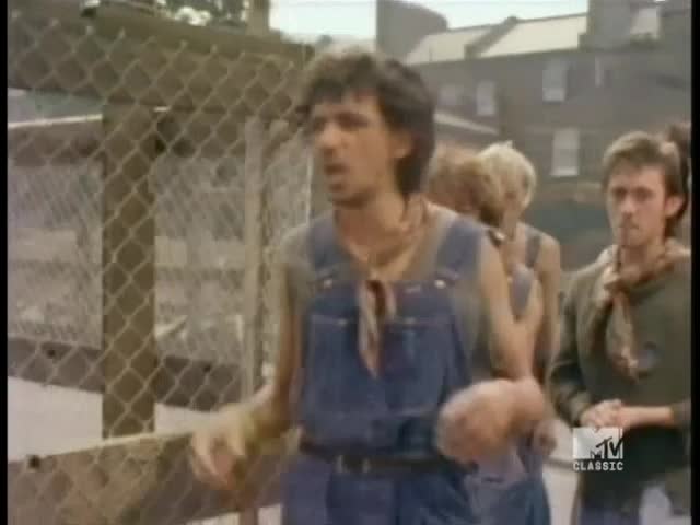 A man in denim overalls gestures with his hands as he walks past a chain-link fence. Behind him, other men in similar outfits follow, the MTV Classic logo visible in the corner.
