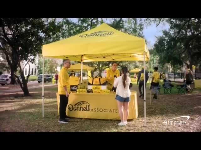 Under a bright yellow tent, people are gathered around a table displaying branded merchandise. A few folks are checking out what's on offer, while others mill about the park.
