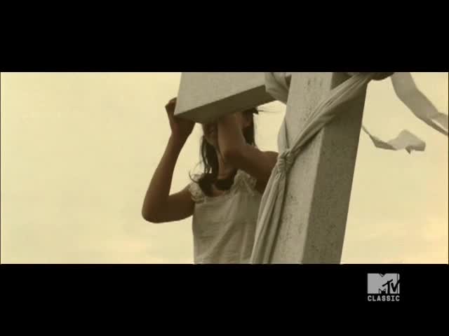 A woman in a white dress struggles to hold a large, stone cross. White fabric flutters in the wind, tied to the cross's arms.
