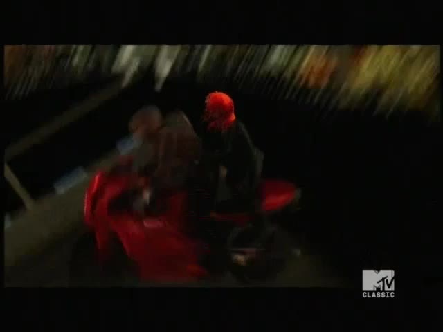 A red motorcycle whips past, carrying two figures. The rider in the back is wearing a bright orange helmet, and the scene is lit by the glow of the MTV Classic logo.
