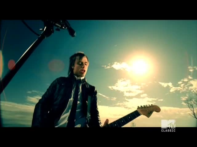 The singer, clad in a leather jacket, is belting out a song, his guitar angled towards the sun. Behind him, the sky is a bright, cloud-streaked canvas, and the MTV Classic logo is visible in the corner.
