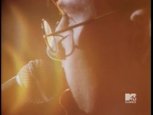 A person with glasses is singing into a microphone, bathed in warm light. The MTV Classic logo is in the corner, and the scene feels like a vintage music video.
