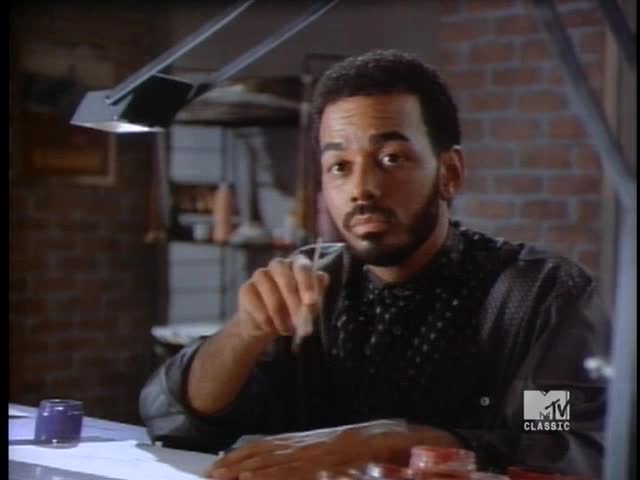 A man with a close-cropped beard holds a brush, looking directly at the camera. He's seated at a table, working under a bright, angled lamp, with the MTV Classic logo visible in the corner.
