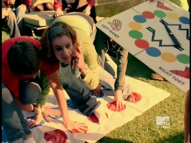 A woman, mid-Twister game, is on the phone, her body contorted to reach the red dot. The spinner holds the game board over the players, directing the next move.
