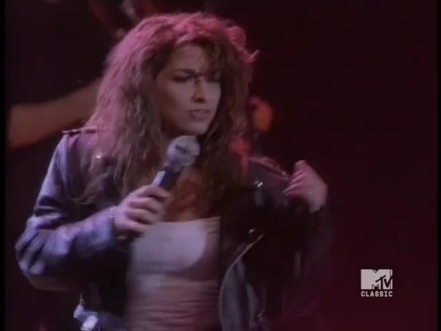 She's holding a microphone, singing with a serious expression. Her leather jacket is open, revealing a white top.
