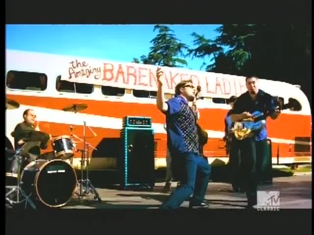 The singer in a patterned shirt is mid-leap, microphone in hand, while the guitarist strums a blue instrument. Behind them, a bus with "The Amazing Barenaked Ladies" painted on it serves as a backdrop.
