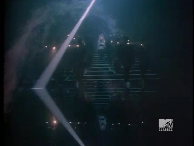 A spotlight pierces the darkness, hitting a mirrored set of stairs. Figures are ascending the steps, bathed in the ethereal glow.
