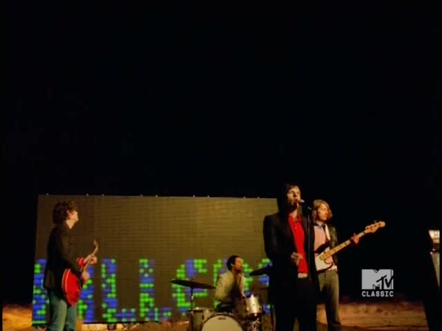 The lead singer, in a red shirt, gestures as he belts out a tune. A guitarist strums next to a backdrop that reads "KILLERS" in pixelated letters, all under the MTV Classic logo.
