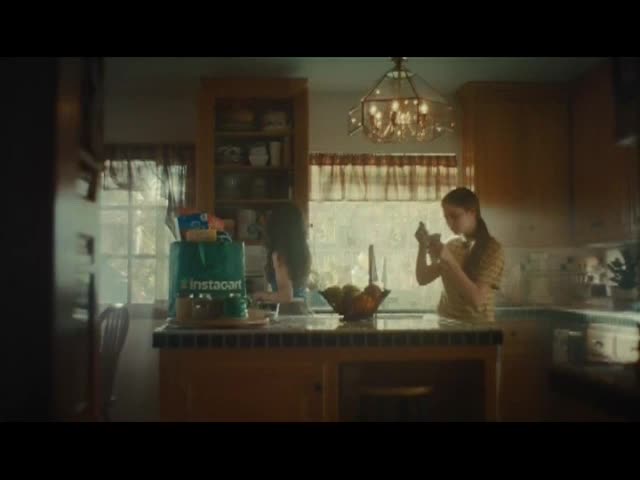 A woman unpacks groceries from an Instacart bag on the counter. Across the kitchen, another young woman eats something, seemingly oblivious.

