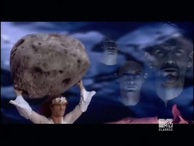 A woman in a white dress holds a massive, pitted rock above her head. Ghostly faces float in the background as the MTV Classic logo appears.
