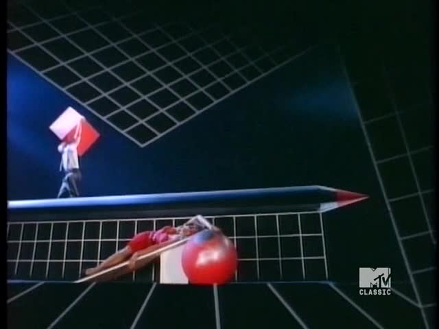 A man in a white shirt and dark pants walks across a narrow beam, balancing a large red and white cube above his head. Below, a woman in a red leotard reclines on a ramp, next to a red ball, all set against a geometric backdrop.
