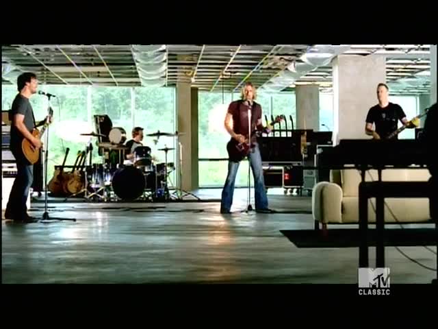 The band is playing in a spacious room with large windows. The singer in the center is holding a guitar, while the drummer keeps the beat.
