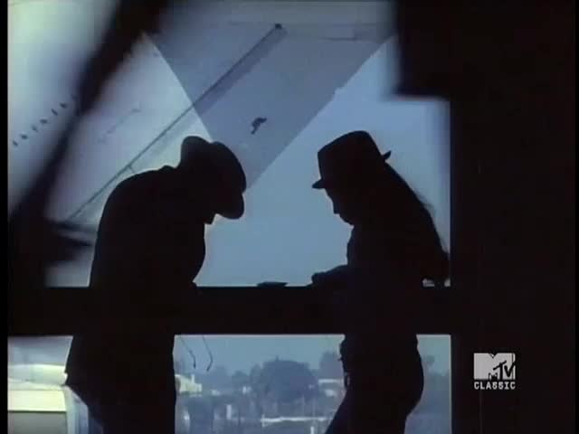 Silhouetted figures stand near a window, their hats casting shadows over their faces. The scene is dark, with the hazy outline of an airplane visible in the background.
