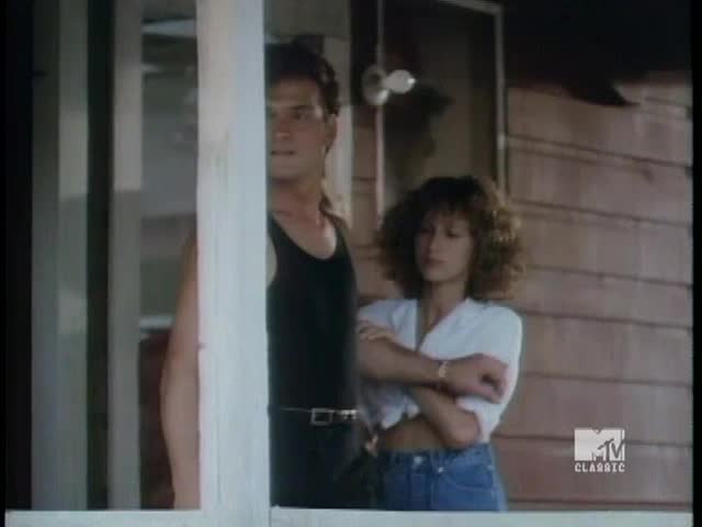 He's peering out from behind a white post, wearing a black tank top and a serious expression. She stands beside him, arms crossed, in a white shirt and jeans, looking off to the side.
