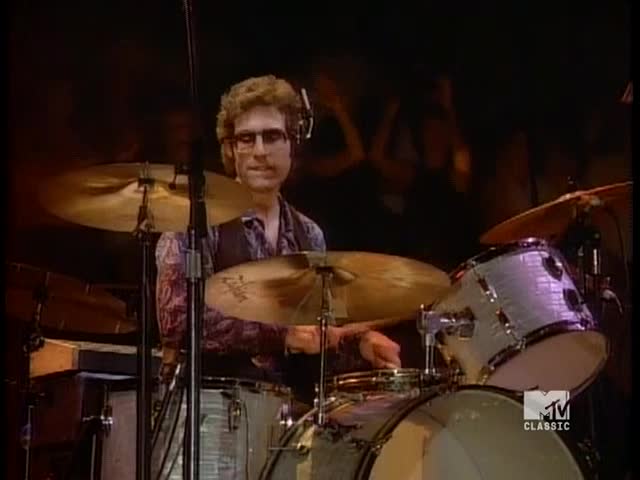 The drummer, wearing glasses and headphones, is focused on his kit, his hands moving quickly.  He's playing on MTV Classic, the cymbals shimmering under the stage lights.
