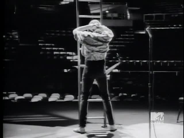 A figure in a patterned jacket stands on the stage, arms raised, leaning against a tall ladder. The black and white image, clearly from an MTV Classic broadcast in the United States, captures a moment of performance.
